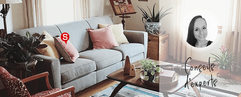 Un salon vintage avec un canapé gris et des coussins moelleux dans les couleurs clairs. Devant le canapé, il y a une table basse en bois foncé. Sur le côté, on voit un fauteil tapissé d'un tissu à motif sixties.